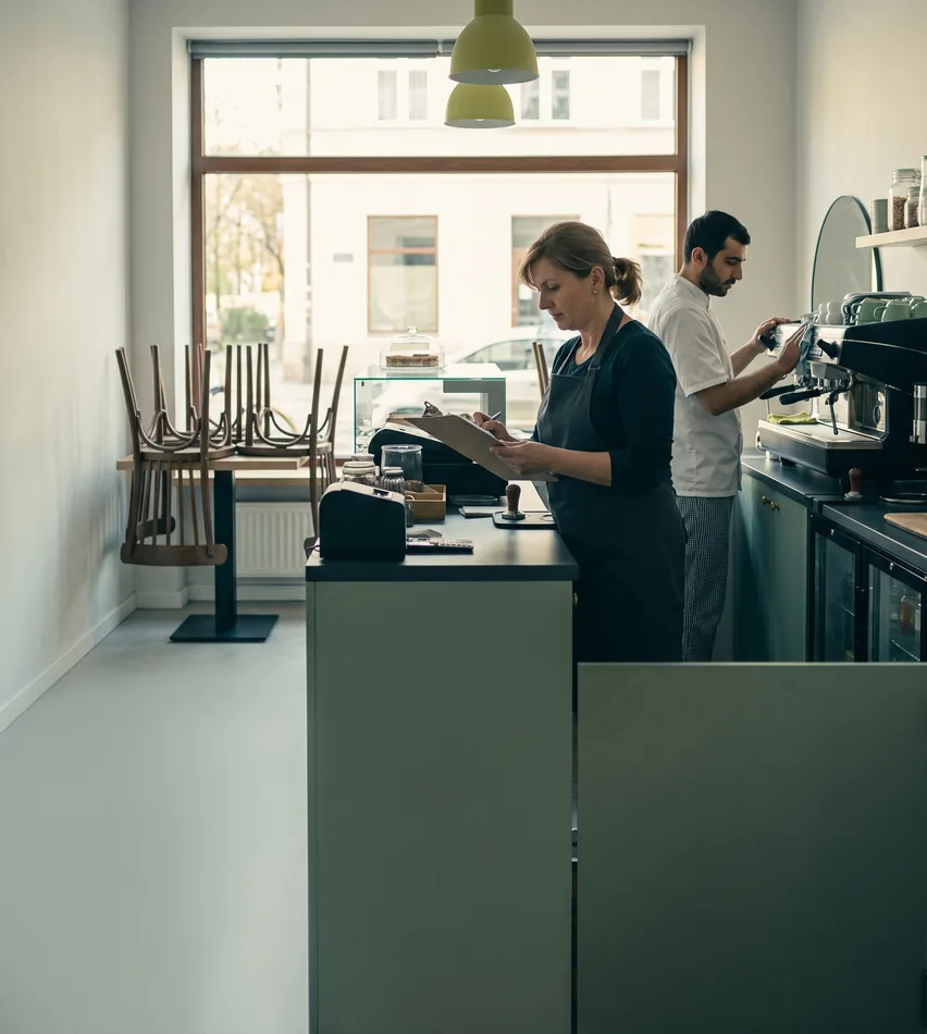 Kobieta z podkładką do pisania przy ladzie kawiarni, barista pracuje przy ekspresie